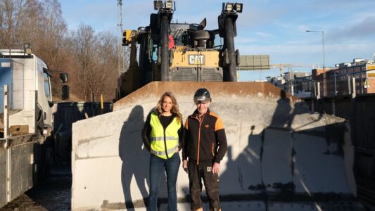 Två personer, en kvinna i reflexväst och en man i säkerhetshjälm och arbetskläder, poserar framför en Caterpillar D8T bulldozer på en byggarbetsplats. Maskinens stora schaktblad är i förgrunden och sträcker sig mot kameran.