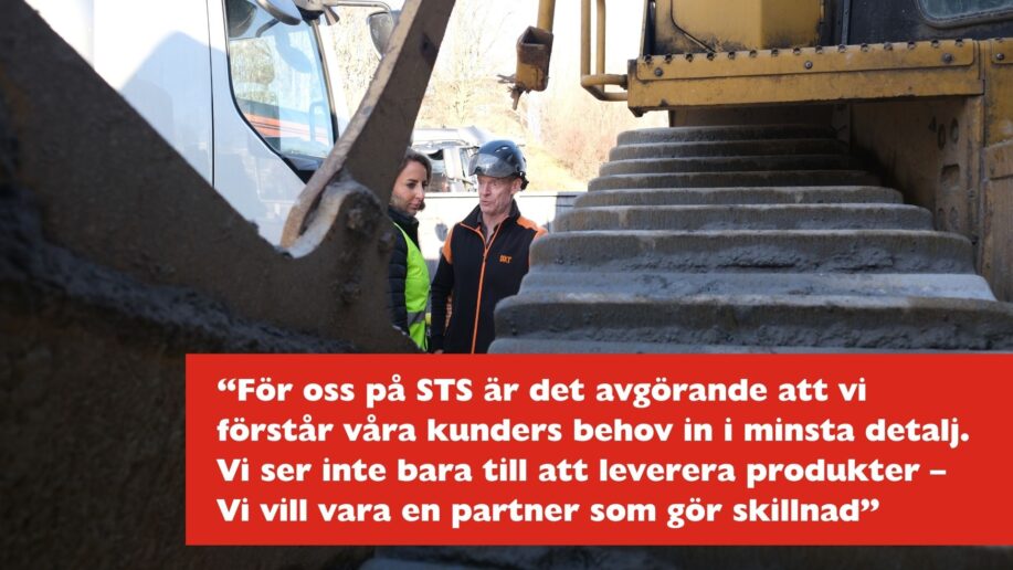 En närbild av Caterpillar D8T:s bandspår på en byggarbetsplats. Två personer i säkerhetskläder, en i en gul reflexväst, står bredvid maskinen och diskuterar arbete, medan en lastbil syns i bakgrunden.